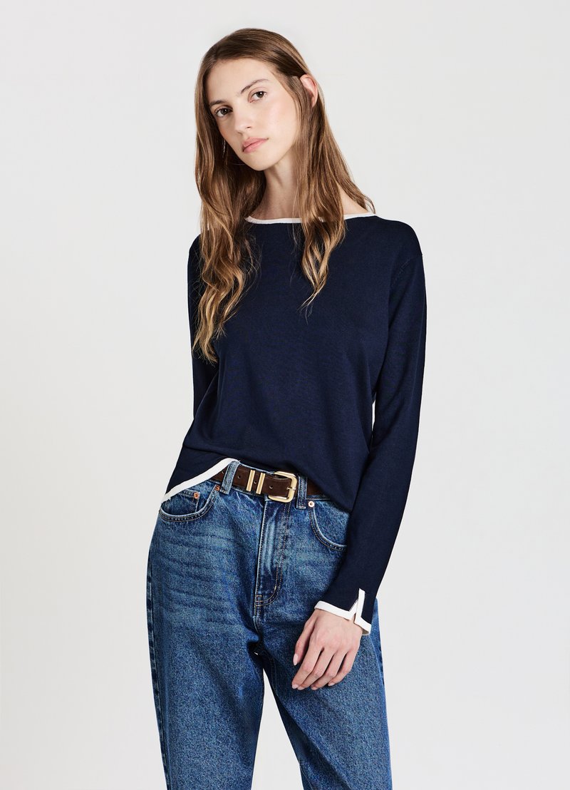 Navyblaues langärmliges Strickoberteil mit weißem Besatz am Ausschnitt und an den Ärmelbündchen, kombiniert mit hochtaillierten blauen Jeans und einem braunen Gürtel.