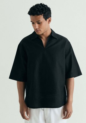 Hombre con el pelo corto y rizado lleva una camisa negra de manga media y cuello, holgada, y pantalones blancos, de pie ante un fondo liso y claro.