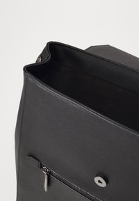 Crna sintetička torba s teksturiranim finišem, koja ima zippered prednju džep i zatvaranje na dugme. Unutrašnjost je obložena tamnom tkaninom.