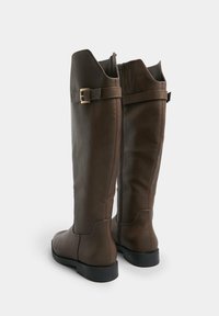 Stivali al ginocchio in pelle marrone con zip sul retro, dotati di un dettaglio con fibbia e suola piatta. Texture liscia, design strutturato.