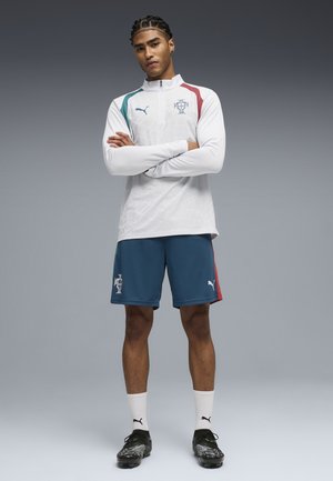 Young man standing with arms crossed wearing Portugal soccer training apparel and black Puma cleats against gray background.