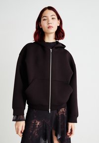 Svart zip-up hettegenser i mykt materiale med stor frontlomme, justerbar hette og ribbede mansjetter. Brukt over en gjennomsiktig mønstret kjole.
