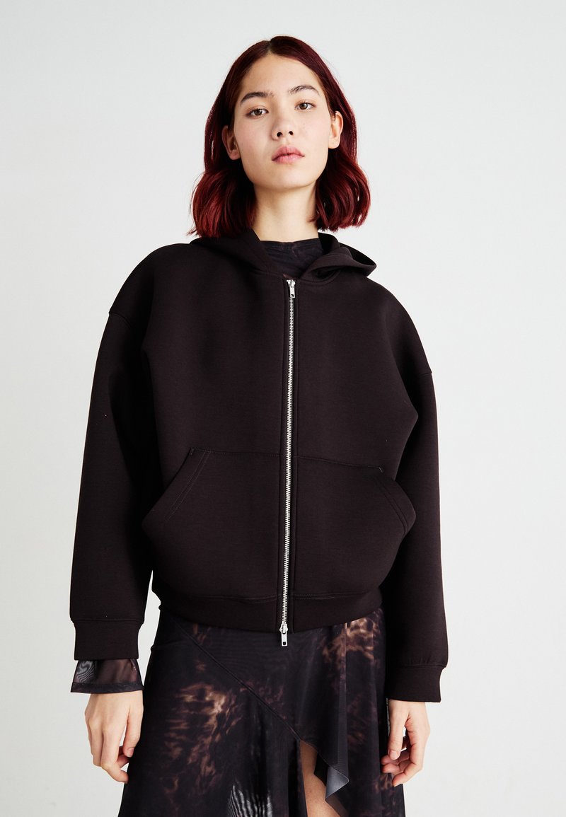 Svart zip-up hettegenser i mykt materiale med stor frontlomme, justerbar hette og ribbede mansjetter. Brukt over en gjennomsiktig mønstret kjole.