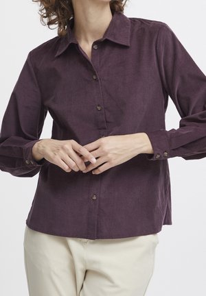 Persona vistiendo una camisa de pana morada oscura de manga larga y abotonada, con pantalones beige, manos entrelazadas frente a la cintura.