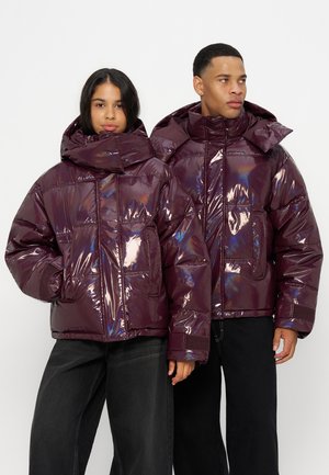 Dvaja ľudia v rovnakých lesklých tmavofialových vatovaných bundách s vysokými goliermi, stojaci proti jednoliatej bielej stene.
