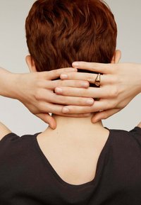 Top negro de manga corta, que deja al descubierto la nuca; manos entrelazadas detrás del cuello. Cabello corto y ondulado de color castaño rojizo. Anillo con diseño dorado texturizado en el dedo.