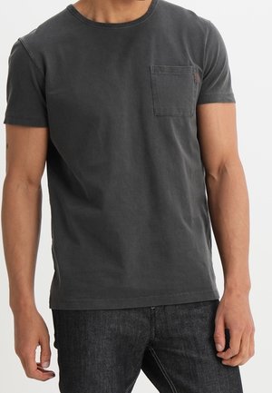 Homme portant un t-shirt noir délavé à manches courtes avec une poche poitrine et un jean foncé sur un fond uni.