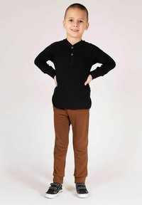 Czarna koszulka henley z długim rękawem i trzema guzikami, zestawiona z brązowymi, dopasowanymi spodniami. Czarne buty z szarymi sznurowadłami, stojąca poza.