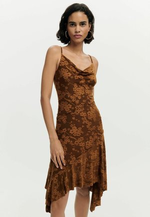 Naine pruunis lillelise mustriga slipkleidis, millel on õhukesed rihmad, asümmeetriline volditud serv ja suured rõngaskõrvarõngad, valge ühevärvilise tausta ees.