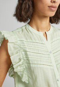 Jasnozielona bluzka z falbanami na rękawach, w pionowe paski, małe zapięcie na guzik oraz delikatne detale z dziurkami na wysokości klatki piersiowej.