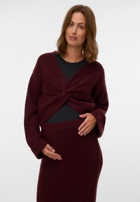 Burgundijska pletena majica s detaljem na prednjem dijelu u obliku twista, u kombinaciji s uskim suknjama u istoj boji. Meka tekstura, opušteni kroj i dugi rukavi.