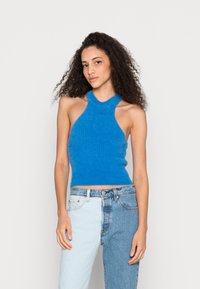 Top halter azul com textura canelada, apresentando laterais abertas. Combinado com jeans que misturam tons de denim claros e escuros. Design simples e ajustado.