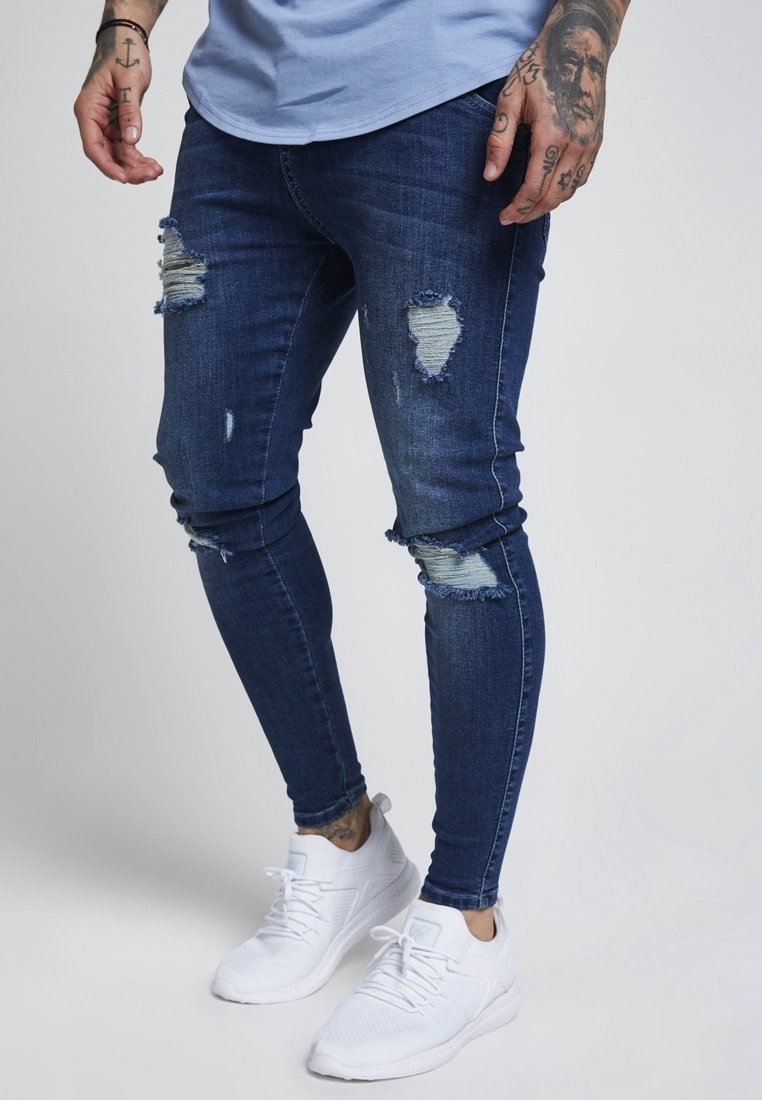 Donkerblauwe gescheurde jeans in een slim fit ontwerp, met versleten details en vervaging. Gecombineerd met witte sportschoenen.