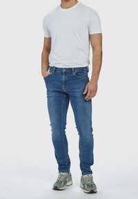 Slim-fit blå denimjeans med medelvattning, fem fickor och dragkedja, kombinerat med en enkel vit t-shirt och grå sneakers.