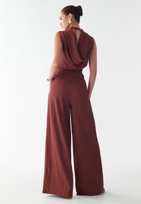 Rostevärviline laiad jalad jumpsuit drapeeritud kaelusega, avatud seljaga ja siledal tekstuuril. Omab kõrge vöökoht ja täispikk disain.