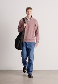 Světle růžová mikina na zip, modré džíny, černé boty a černá kabelka přes rameno. Model kráčí proti jednobarevnému bílému pozadí.