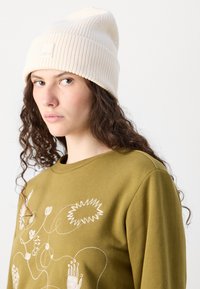Bonnet tricoté blanc à côtes avec un revers, associé à un sweat-shirt vert arborant des motifs abstraits brodés en beige et blanc.