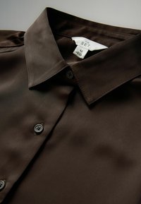 Mørkebrunt skjorte med knaplukning og klassisk krave samt synlig mærke med teksten "THE SET", størrelse M, fremstillet i Bangladesh.