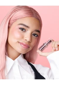Lipstick in a clear tube with a metallic silver base, held by a person with pink hair, wearing a white collared shirt against a pink background.