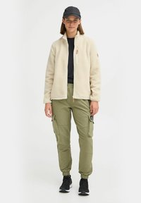 Veste en polaire crème, devant zippé, poches latérales. Pantalons cargo olive vert avec chevilles élastiques, plusieurs poches, chaussures noires, casquette noire.