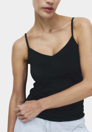 Vrouw draagt een zwart spaghettihemd met een V-hals, leunt ontspannen met één hand op haar taille, tegen een witte achtergrond.