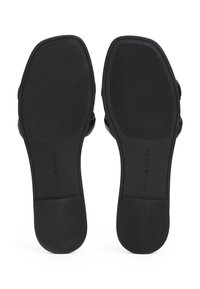 Paire de sandales plates noires vues par la semelle, montrant des orteils carrés et des semelles en caoutchouc texturées avec le nom de la marque en creux.