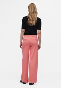 Pantalones de pierna ancha rosa con una cinturilla elástica, dos bolsillos traseros y una textura suave. El conjunto se completa con una blusa negra de canalé y zapatos negros.