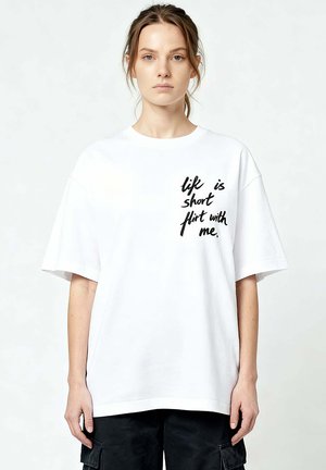 Giovane donna con capelli castano chiaro raccolti in una coda di cavallo morbida, indossa una maglietta bianca oversize con testo nero "la vita è breve, provocami" e pantaloni neri.