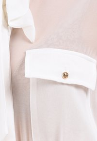 Hvid bluse med en gennemsigtig tekstur, der har en brystlomme prydet med en guldknap. Blødt, let stof med flæse-detaljer.