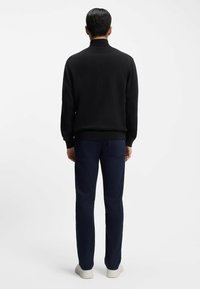 Mężczyzna stojący tyłem, ubrany w czarną koszulkę z długim rękawem, granatowe spodnie i białe trampki na jednolitym białym tle.