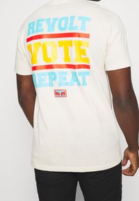 Krémové bavlněné tričko s velkým, barevným textem: "REVOLTUJ," "HLASUJ" a "OPAKUJ" v modré, žluté a červené. Malé logo dole. Krátké rukávy.