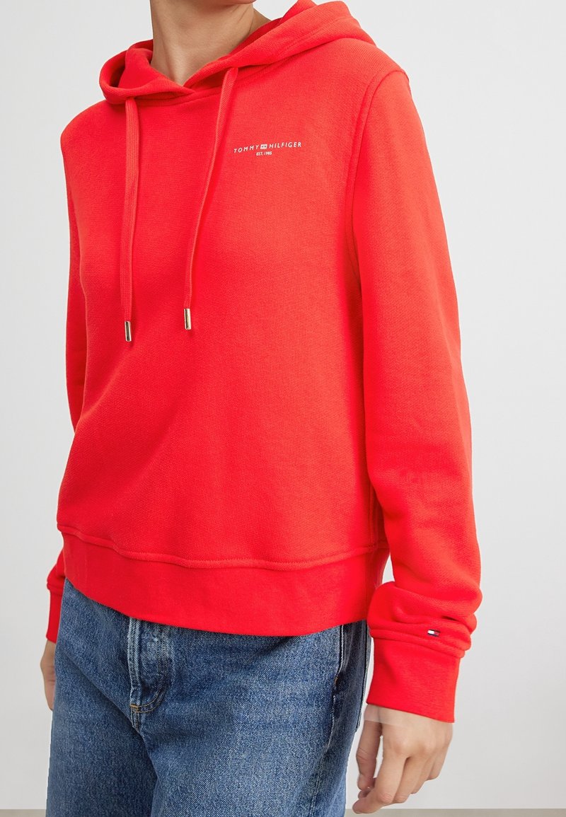 Personne portant un sweat à capuche Tommy Hilfiger rouge vif avec des cordons et un jean en denim bleu, montrée du cou aux hanches.