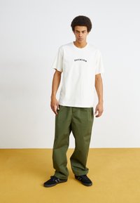 White cotton t-shirt with "DICKIES" text, paired with loose green pants and black sneakers, standing on a yellow floor.