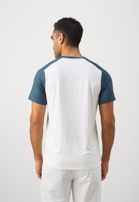 Herre treningstrøye med hvit kropp og teal raglan-ermer. Laget av lett, teksturert stoff med rund hals og korte ermer.