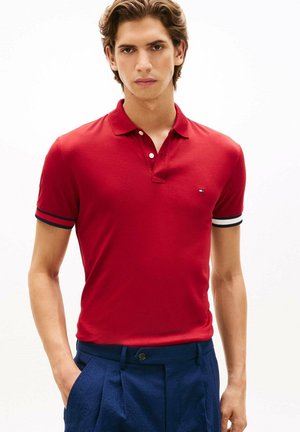 Giovane uomo con indosso una polo rossa con polsini a righe e pantaloni blu, in piedi con una mano in tasca su sfondo bianco.