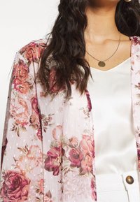 Žena v květované průsvitné růžové blůze přes bílém saténovém topu, s kulatým náhrdelníkem a bílými kalhotami s knoflíkovým poutkem.