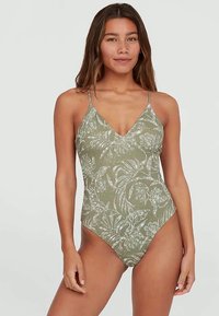 Olíva zöld, fehér virágmintás, vékony pántos egyrészes fürdőruhát viselő nő, sima háttér előtt állva.