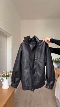 Giubbotto di pelle nera con un design strutturato, dotato di un alto colletto, bottoni a pressione e cuciture diagonali sulla parte anteriore.