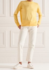 Person med en gul långärmad tröja, vita byxor, vita sneakers och ett enkelt halsband, som står inomhus mot en panelvägg.