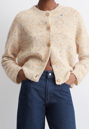 Osoba ubrana w kremowy sweter na guziki z drobnym wzorem, wykończony prążkowanymi krawędziami, oraz niebieskie, wysokowalne dżinsy, z rękami schowanymi w kieszeniach.