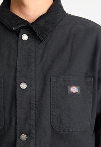 Black Dickies work shirt with corduroy collar, metal buttons, and logo patch on the left chest pocket, worn by a person.