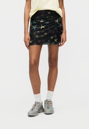 Άτομο που φοράει μαύρη μίνι φούστα με πολύχρωμα σχέδια αστεία, λευκές κάλτσες, γκρι αθλητικά παπούτσια και ανοιχτό κίτρινο επάνω μέρος.