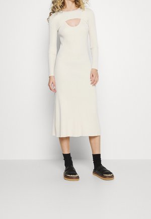 Nainen, joka pukeutunut kermaan vivahtavaan midimittaiseen ribbineulottuun mekkoon, jossa on pitkät hihat ja rinnan avoin leikkaus, yhdistettynä mustiin sukkiin ja avoimilla varpailla varustettuihin sandaaleihin, joissa on korkkipohjat.