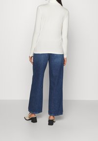 Langärmliger weißer Rollkragenpullover kombiniert mit weit geschnittenen blauen Jeans aus Denim, die einen lässigen Schnitt und einen rohen Saum haben. Geschlossene, hochhackige Schuhe.