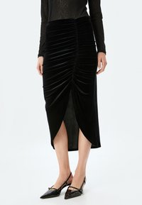 Zwarte fluwelen rok met gerimpelde details en een voorkapsluiting, knielengte, gladde textuur. Gemaakt in combinatie met zwarte schoenen met een spitse neus.