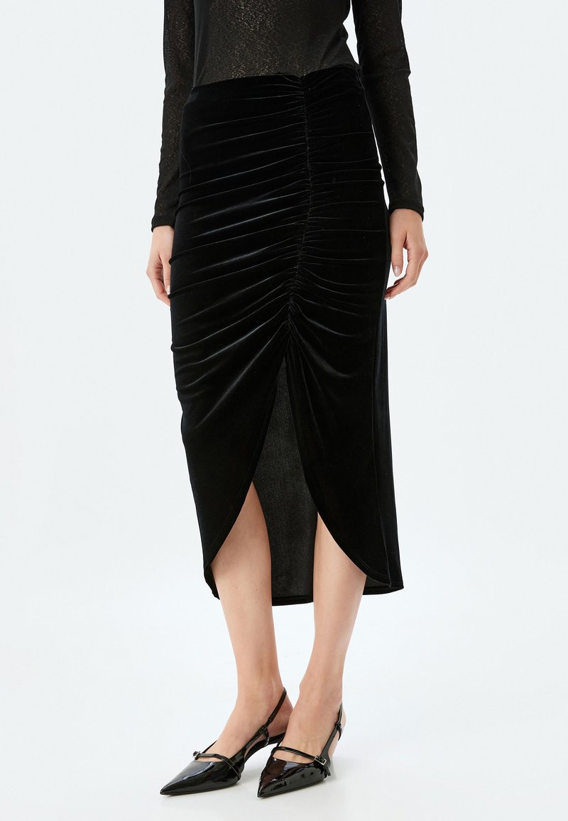 Zwarte fluwelen rok met gerimpelde details en een voorkapsluiting, knielengte, gladde textuur. Gemaakt in combinatie met zwarte schoenen met een spitse neus.
