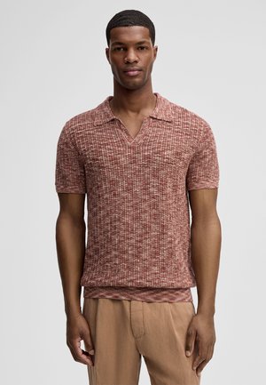 Uomo che indossa una polo a maniche corte in maglia testurizzata in tonalità miste di rosso e pantaloni beige, in piedi davanti a uno sfondo grigio chiaro uniforme.