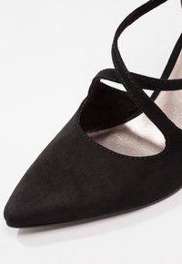 Chaussure femme noire à bout pointu avec des brides croisées à la cheville et une texture douce rappelant le daim.