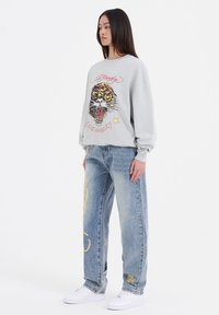 Lichtgrijze sweatshirts met een tijgerafbeelding en tekst, gecombineerd met lichtblauwe baggy jeans met kleurrijke borduursels en witte sneakers.