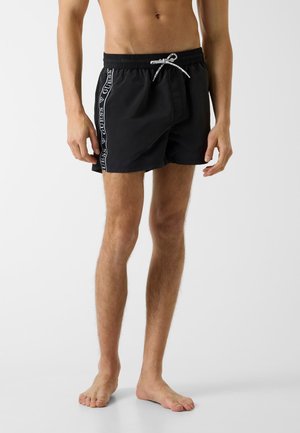 Mann trägt schwarze Badeshorts mit weißem Kordelzug und weißem "GUESS"-Logo an den Seitennähten, steht barfuß vor weißem Hintergrund.